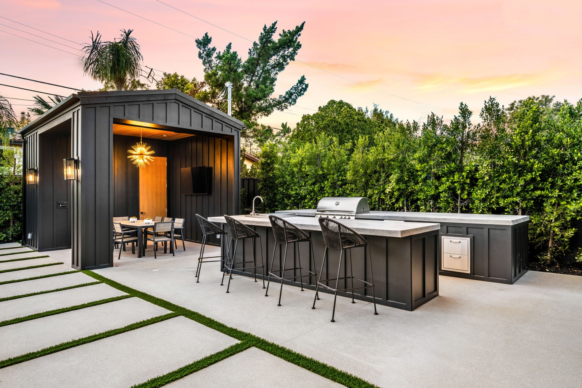 Modern outdoor kitchen with a dining area, surrounded by lush greenery. Barstools line the counter under a pastel sunset sky.