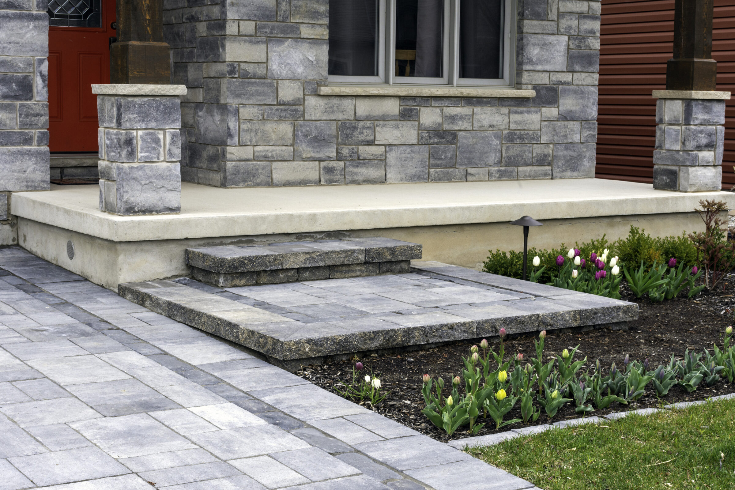 A stone porch with a red door, bordered by blooming tulips in a landscaped garden, leads to a neatly paved pathway.