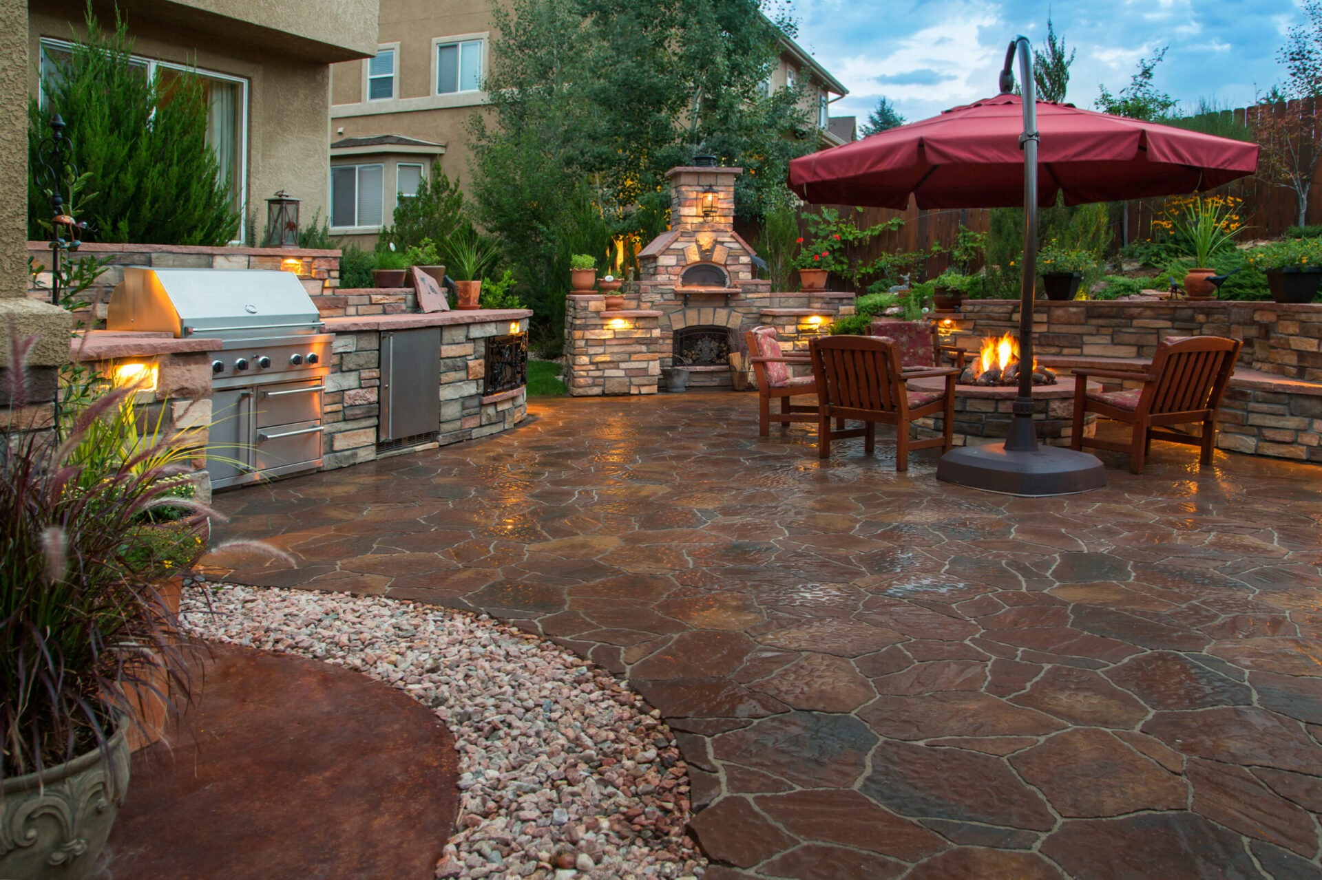 Outdoor patio features a stone kitchen, fireplace, seating area, and umbrella in a landscaped yard with plants and soft lighting.