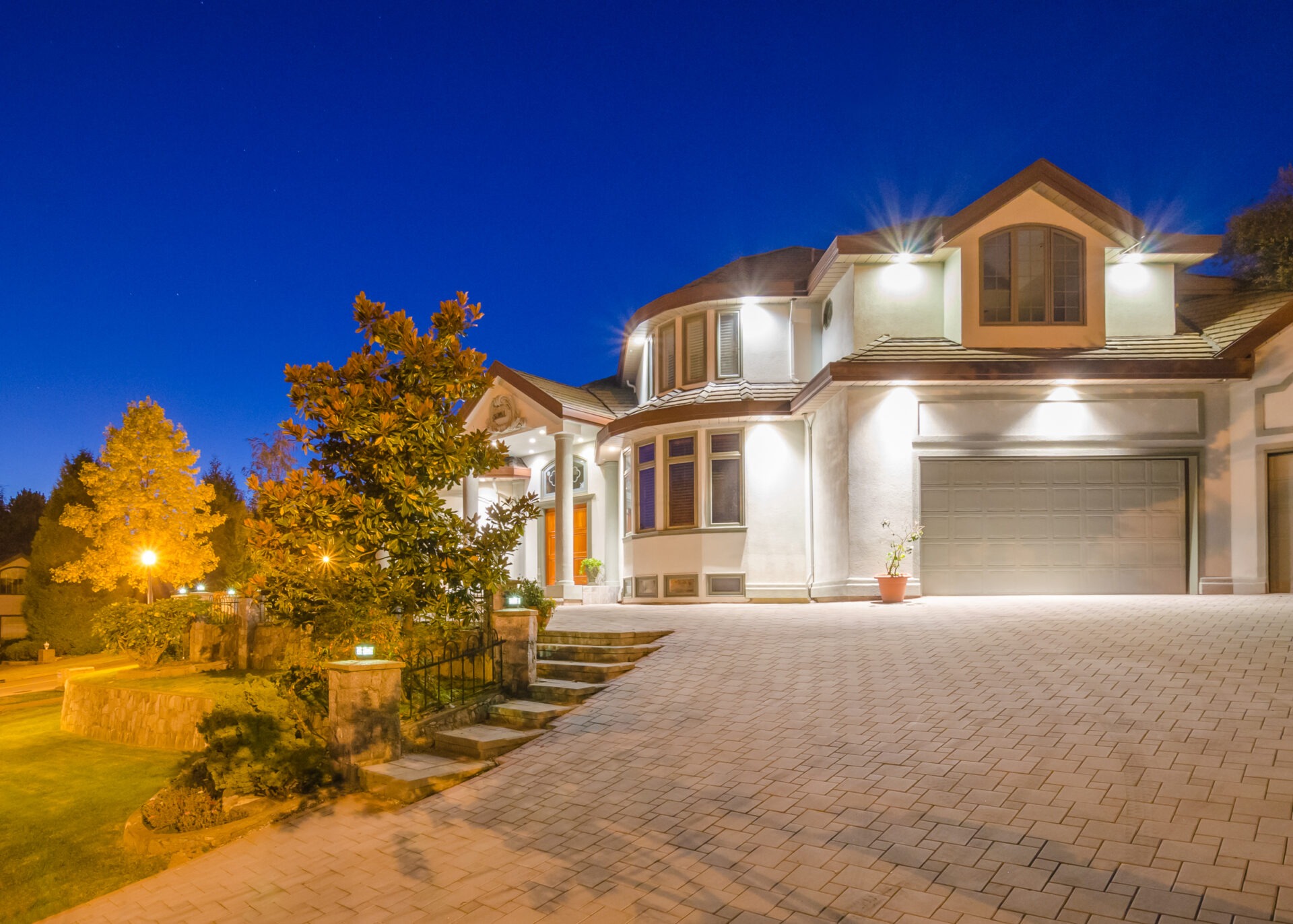 Elegant suburban house at night, well-lit exterior with large driveway, surrounded by trees and landscaped garden, under a clear, deep blue sky.