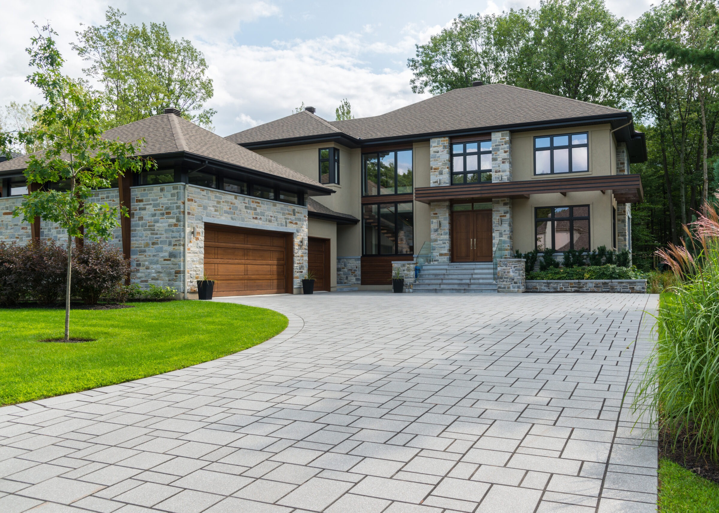 Modern two-story house with a stone facade, large windows, and a spacious paved driveway, surrounded by lush greenery on a sunny day.