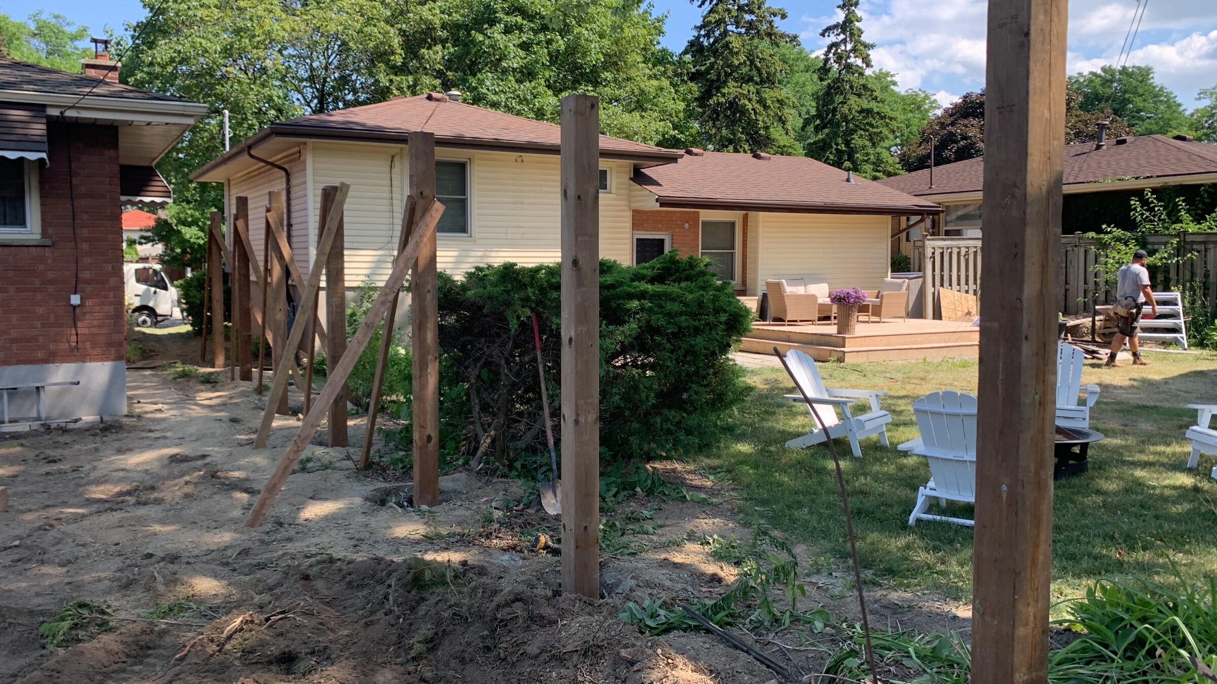 Backyard scene with a partially constructed fence, wooden deck, patio furniture, and person carrying materials. Trees and blue sky in background.
