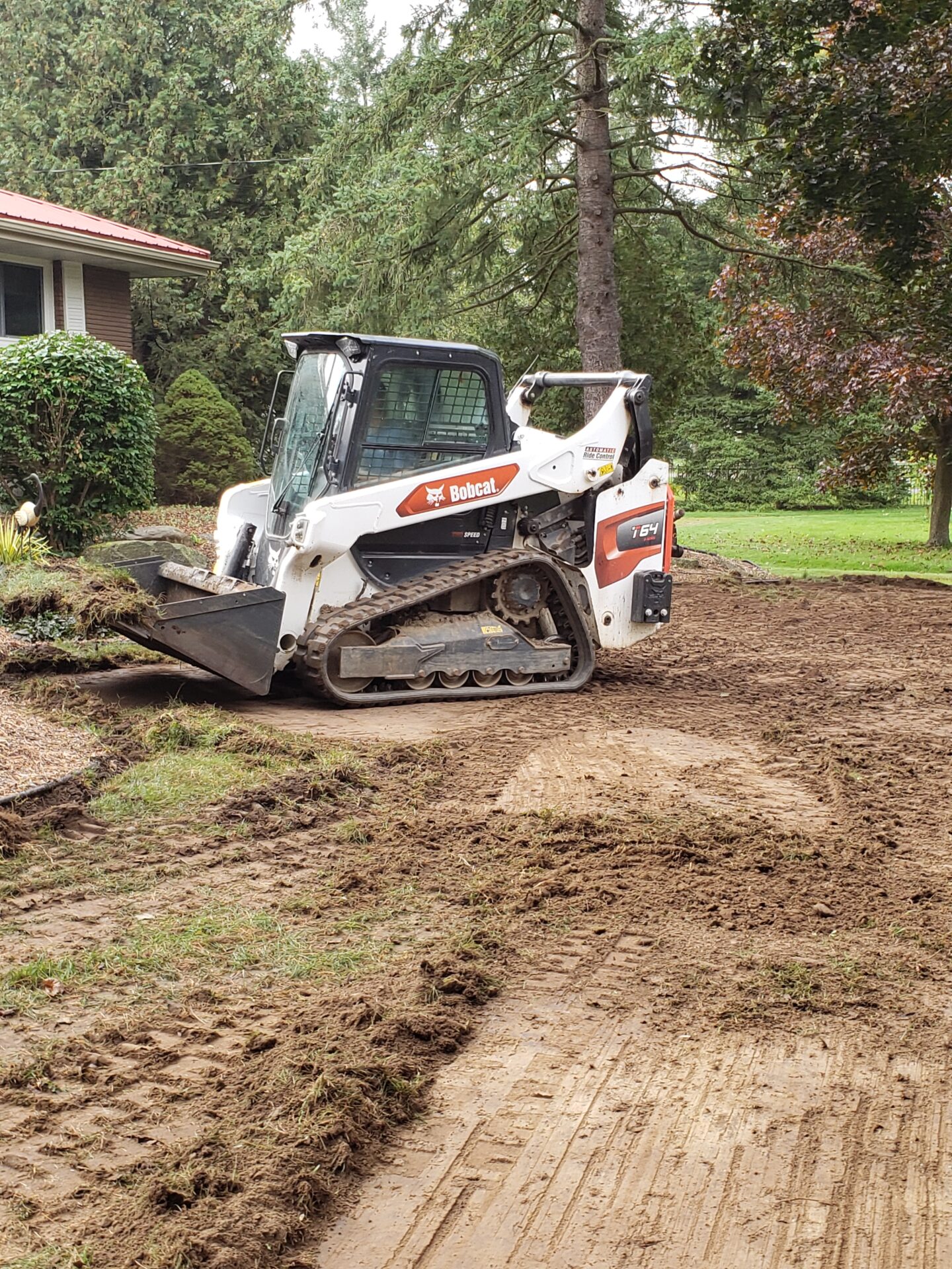 A Bobcat machine is clearing dirt in a garden. Trees and a house with a red roof are visible in the background.