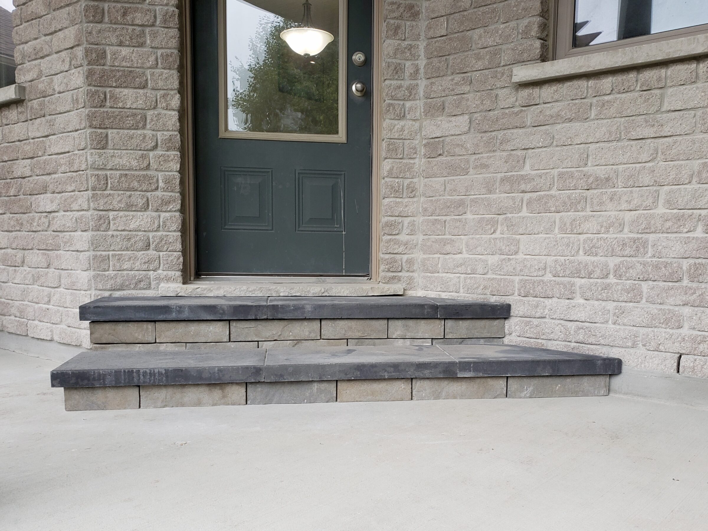 A stone porch with two concrete steps leading to a dark green door with a glass window and visible interior light.
