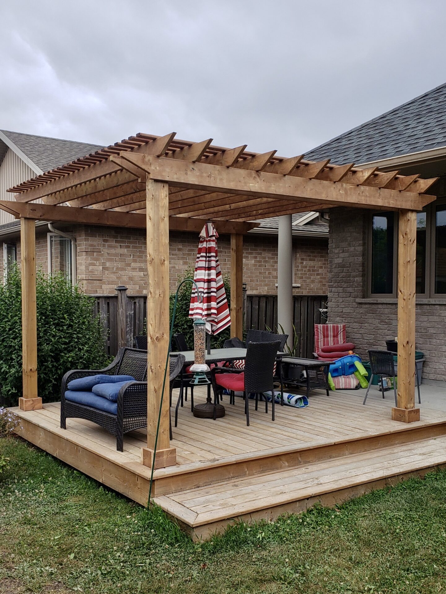 A wooden pergola with seating and a striped umbrella on a backyard deck, adjacent to a brick house, under a cloudy sky.