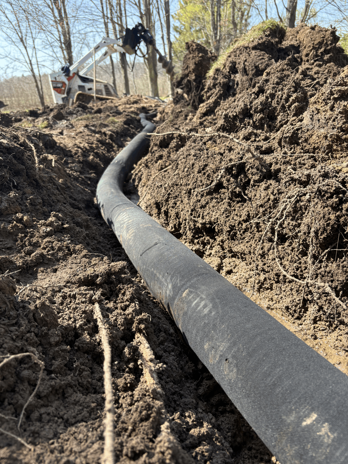 A construction scene showing a large pipe being installed in a trench as an excavator operates amidst a backdrop of trees.
