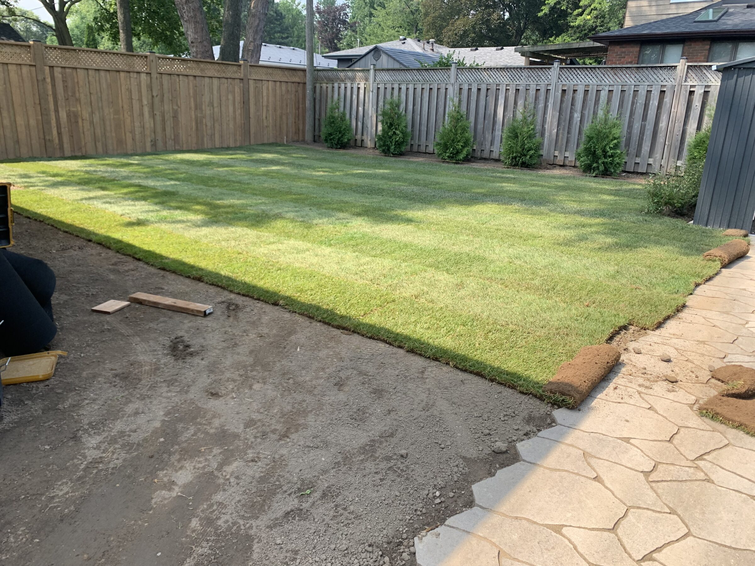 A fenced backyard with newly laid sod, partially finished. Tools and a person's leg are visible near a stone patio.