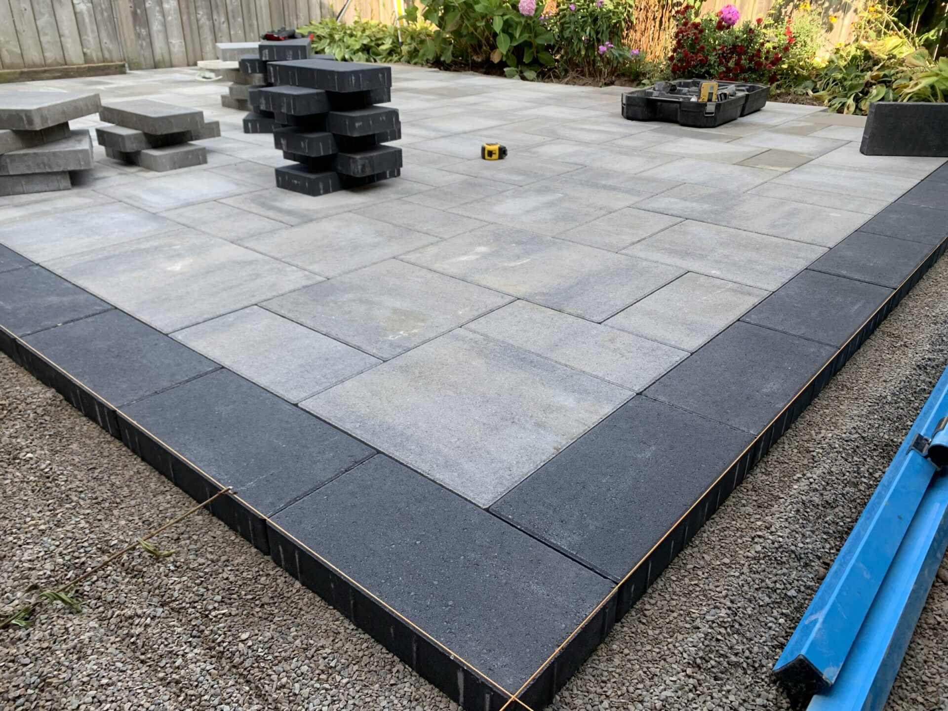 A freshly laid patio with gray and black tiles, surrounded by gravel, under construction with stacked pavers and nearby gardening tools.