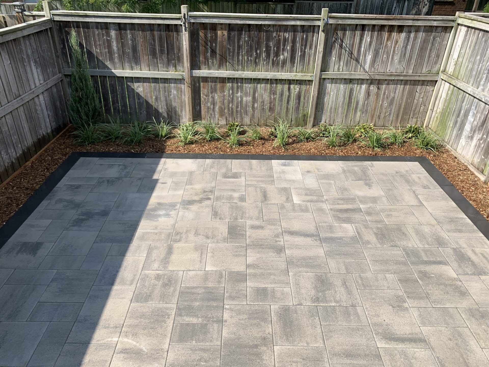 A fenced backyard features a neatly arranged stone patio with a bordering garden bed, planted with small green shrubs and wood mulch.