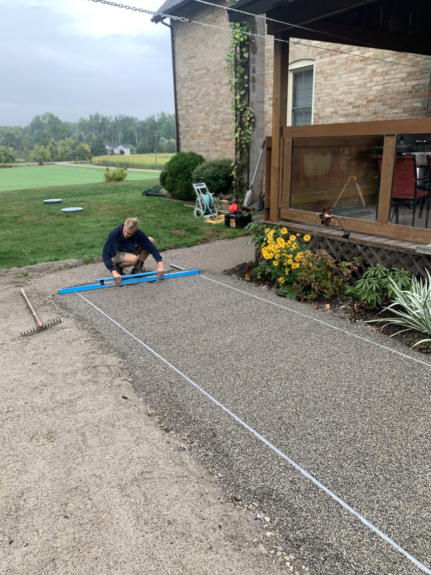 A person adjusts gravel with a tool near a brick house, surrounded by garden plants and grassy landscape. Overcast sky in the background.