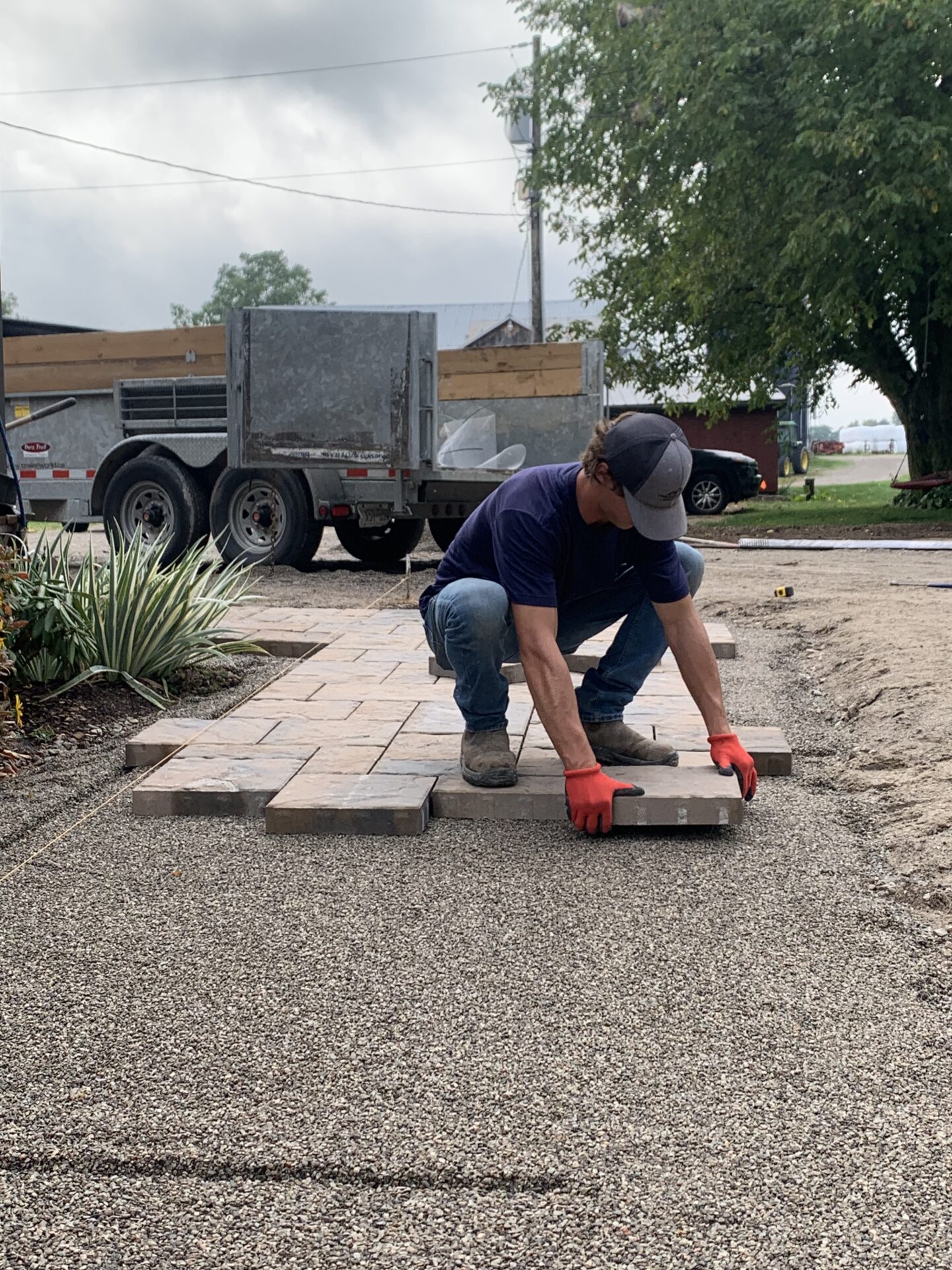 A person wearing gloves lays paving stones in a garden. A trailer and parked car are visible nearby, under a cloudy sky.
