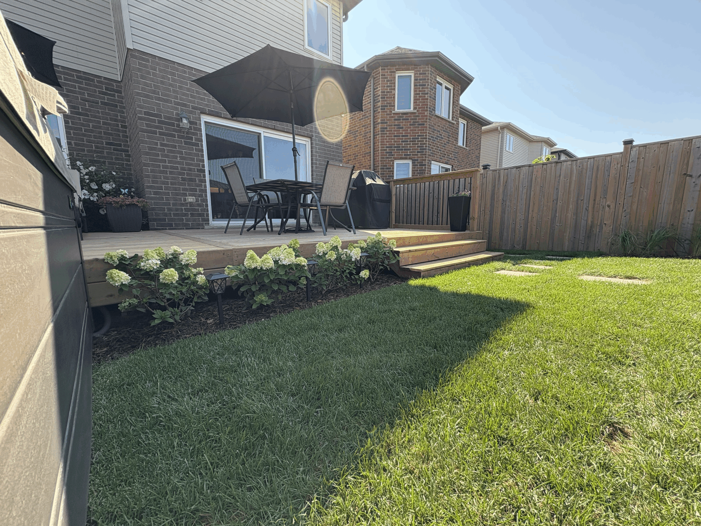 A backyard features a wooden deck with patio furniture, black umbrella, blooming flowers, wooden fence, and neatly trimmed grass under a clear blue sky.