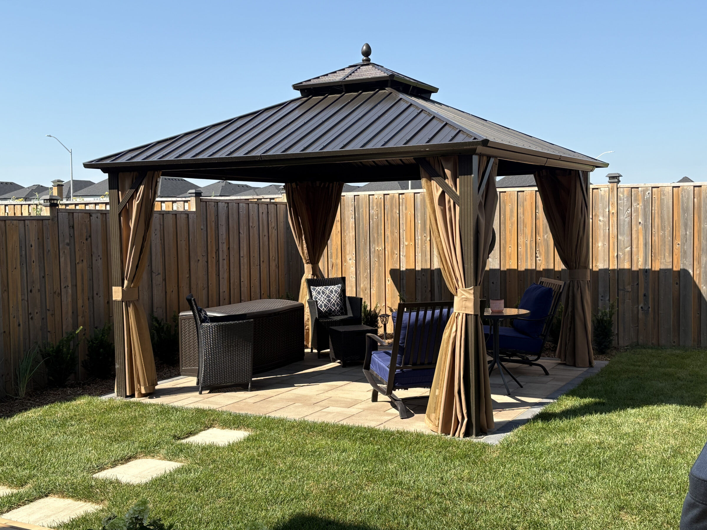 A backyard gazebo with brown curtains and outdoor seating on a grass-lawn patio, surrounded by a wooden fence under a clear blue sky.