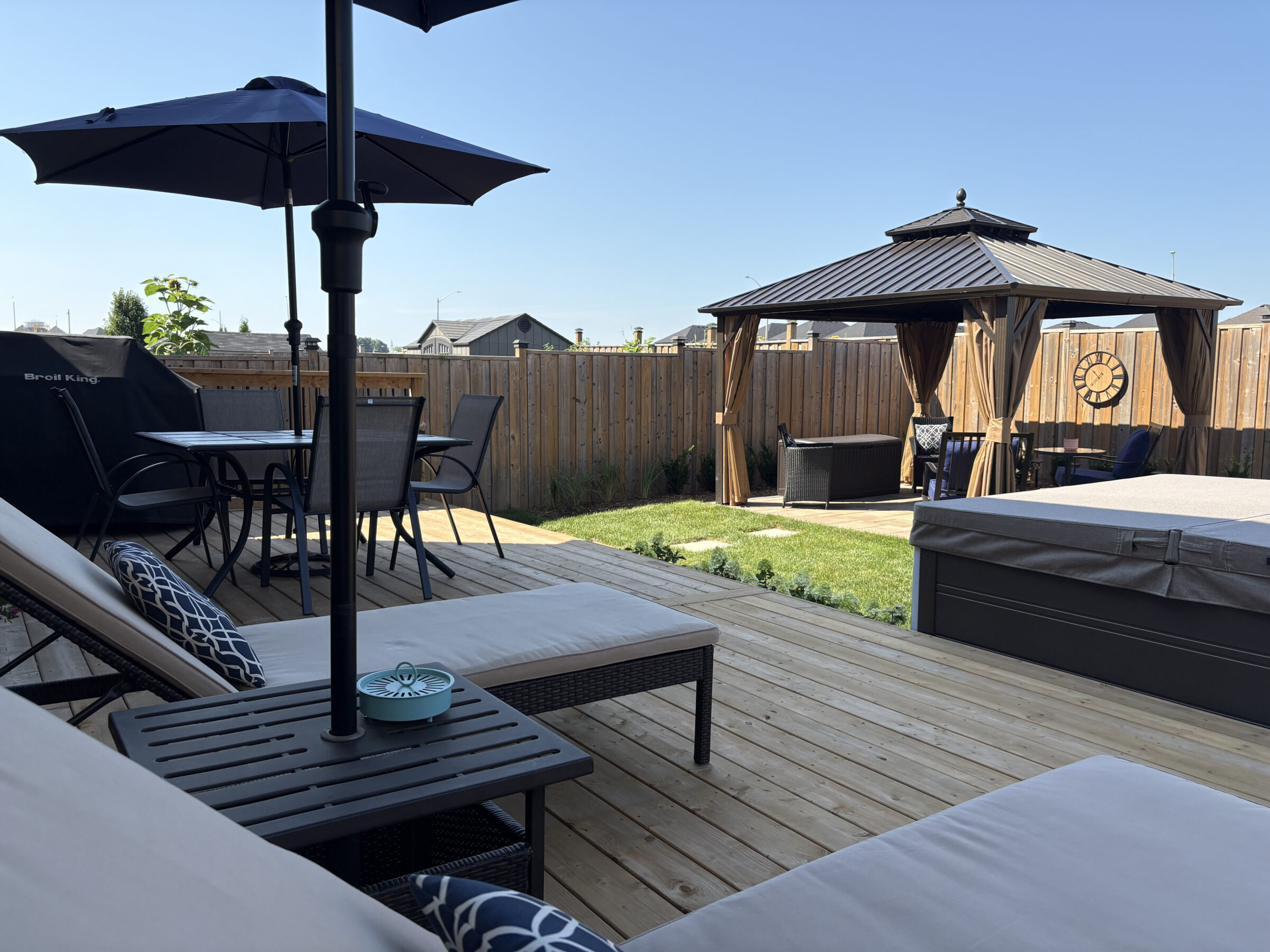 A backyard with a wooden deck, patio furniture, umbrella, gazebo, and hot tub. Fenced-in area under clear blue sky.