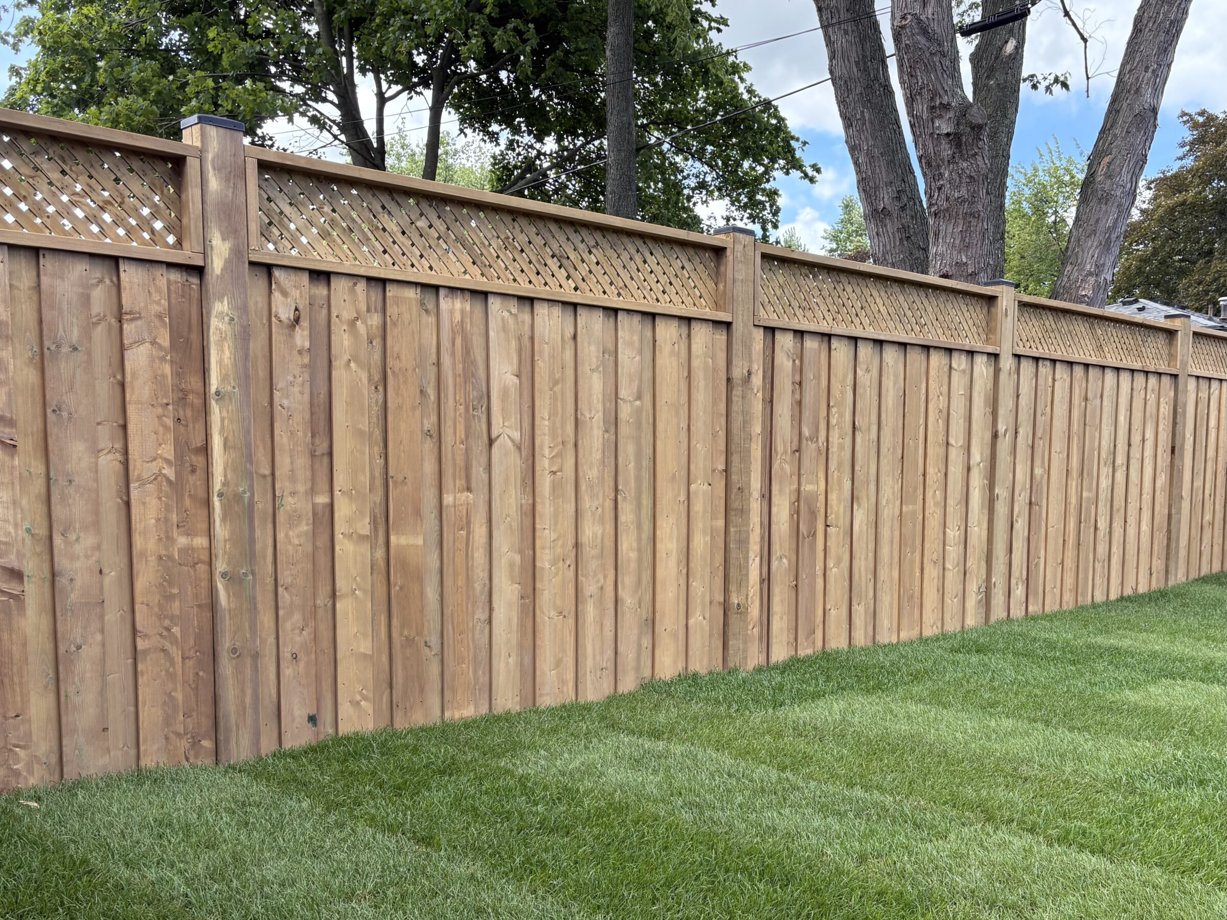 Wooden fence with lattice top stands on neatly trimmed green grass, surrounded by trees under a partly cloudy sky.
