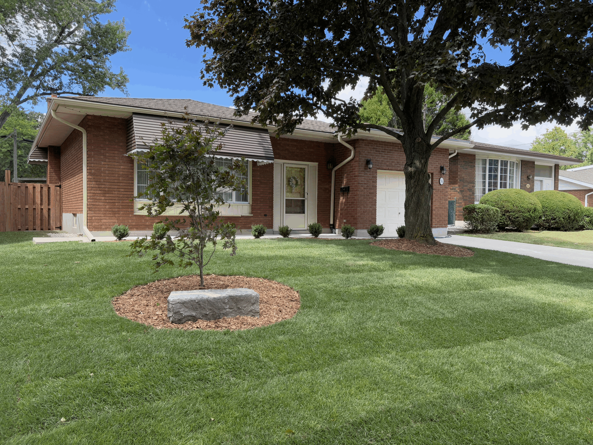 A well-maintained brick house with a lawn, tree, and driveway. The yard is landscaped with bushes and a small tree in mulch.