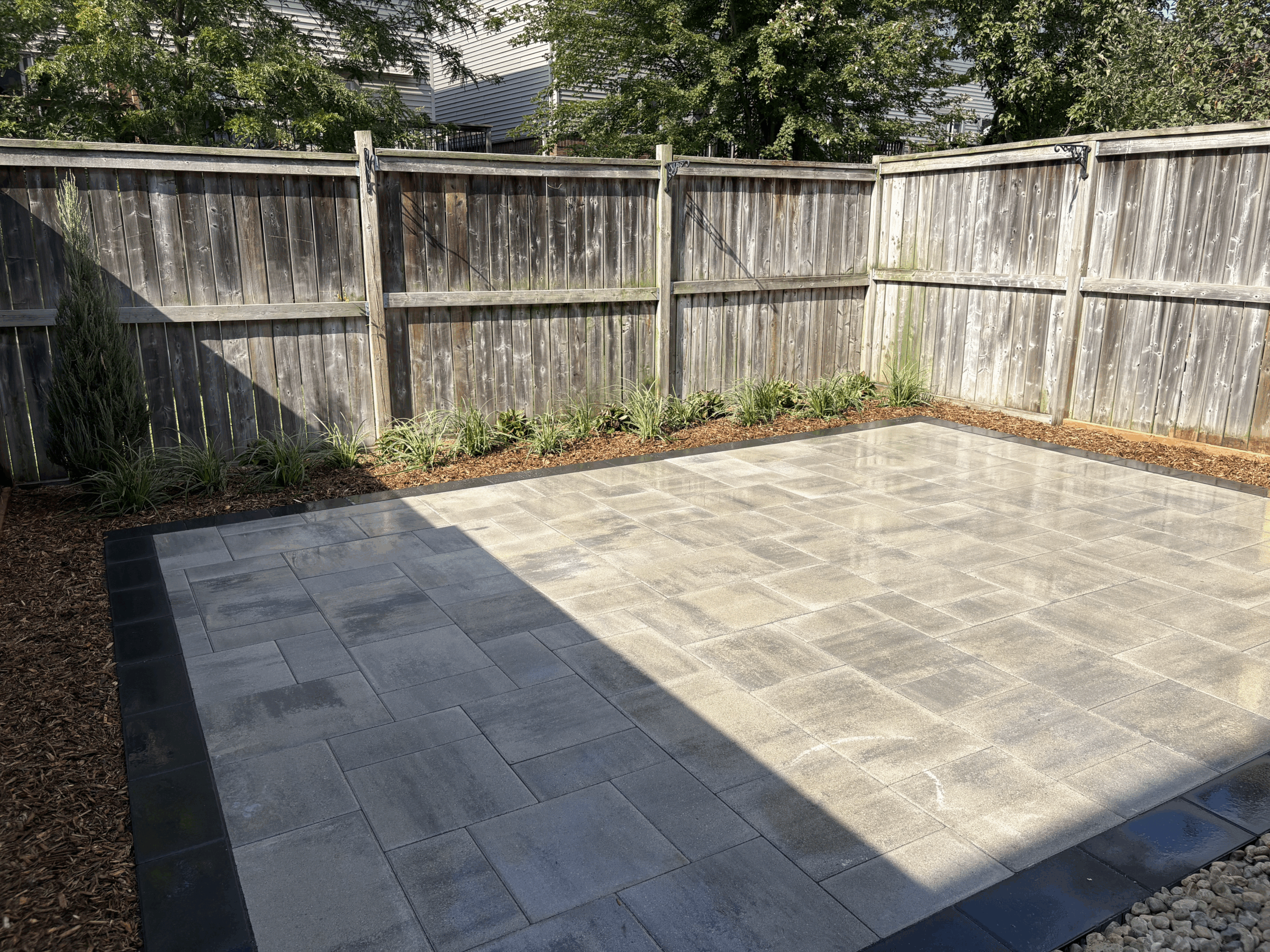 A fenced backyard features a neatly paved patio, surrounded by small plants and bark mulch, under a clear sky.