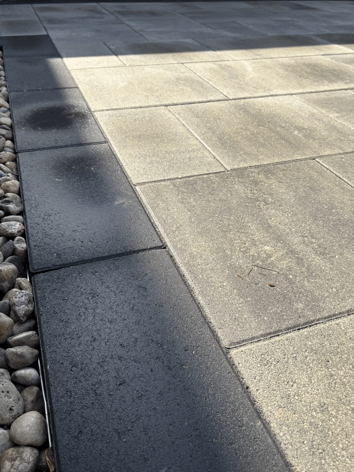 Sunlit patio with light and dark stone tiles, bordered by small river rocks. Peaceful, geometric arrangement enhances the outdoor space's modern design.