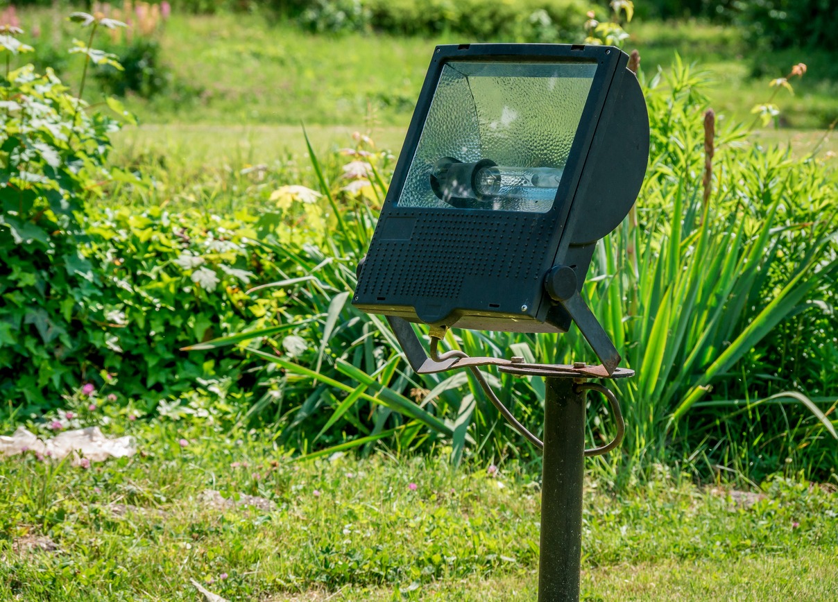 A large spotlight is mounted on a pole in a lush, green garden setting with various plants around.