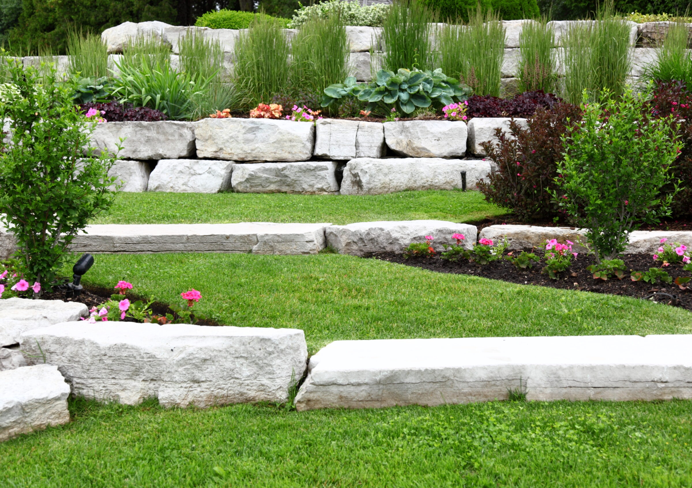 Tiered garden with lush greenery and colorful flowers, bordered by large stone blocks, creating a tranquil and structured landscape design.