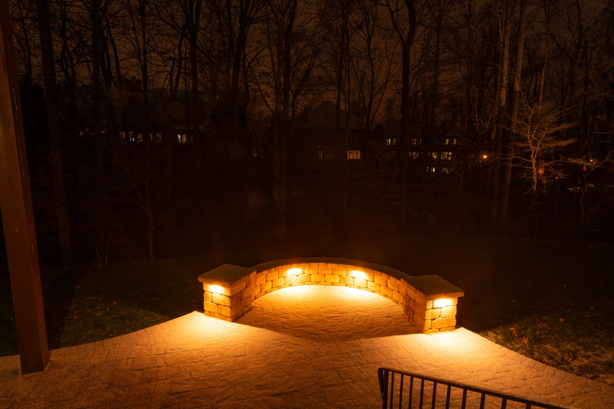 A dimly lit brick wall and pathway, surrounded by leafless trees at night, with illuminated windows visible through the forest.
